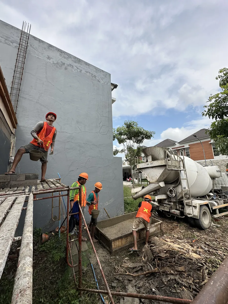 Tukang dengan helm dan rompi keselamatan mengangkut adukan beton dari truk molen saat pengecoran struktur rumah